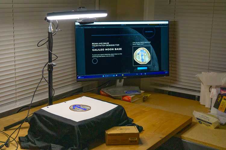 Image of prototype station camera rig, with a raised table at the foot of a small tripod holding up a webcam and selfie light ring, with Moonshot software running on a display behind the rig, and the drawing on the station's table clearly visible on the screen.