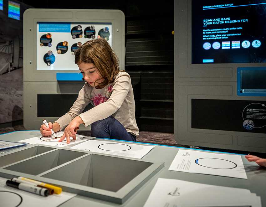 A child drawing at a table. Behind them is a mobile kiosk with a touch screen featuring the Moonshot: Design a Mission Patch software.