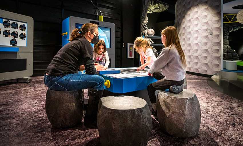 Visitors sit around drawing table at Moonshot Museum. Among them is an instructor speaking with three small children drawing.