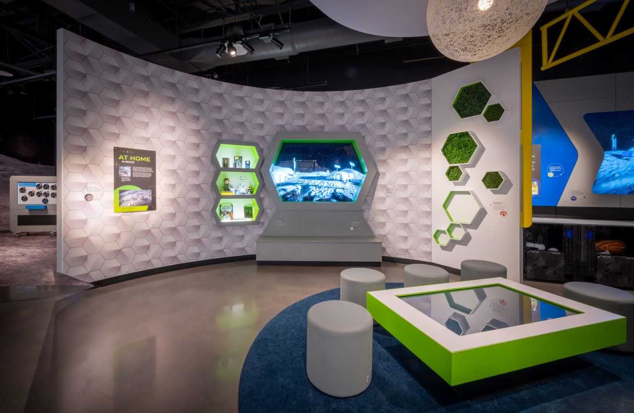 Image of the Space Debates interactive without visitors: A knee-height table with rectangular, green and white casing stands in front of a screen with hexagonal green and white casing, in a wall also featuring a bookshelf with plants.