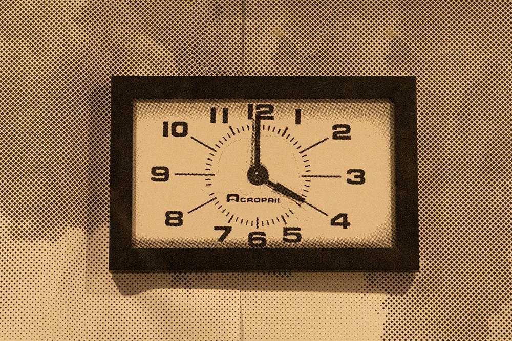 Picture of a framed image of a clock, hung from a wall wallpapered with the images of smokey clouds. The clock's outside body is square, with only two hands tracing the circle the numbers in the center. Printed underneath the numbers, partially obscured, reads the word 'Acropa.' The time reads 4 p.m.
