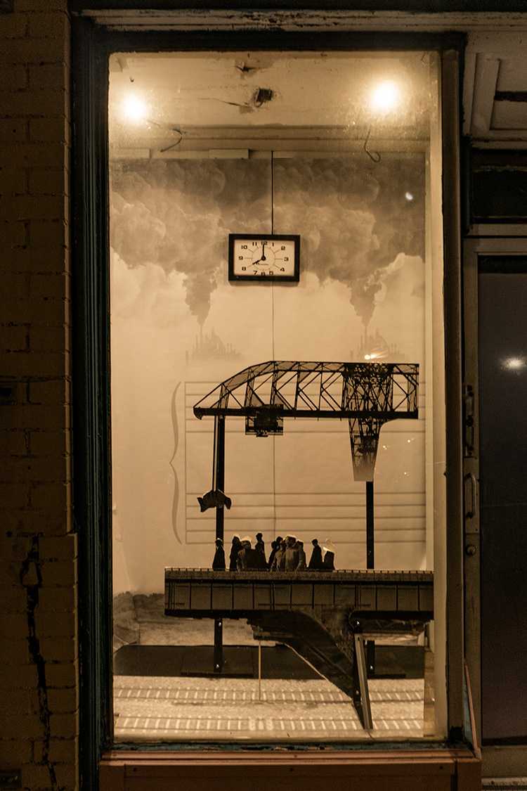 Picture of one bay window, from the front. Inside the windows stands two cutouts of foamcore, one printed with the image of an ore crane - which looks like a truss bridge with big metal shovels hanging by cables underneath, it's mechanical jaw open as if about to pick something heavy up - and the other printed with a road bridge with pedestrians walking across it. Behind these cutouts, the walls of the bay window are papered with wallpaper, which shows clouds and the beginning of musical staff, both treble and bass. A framed picture of a clock hangs in the center of the back wall of each window. Its time reads 8 a.m.